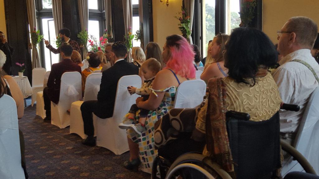 guests at a wedding, congregation seated on white chairs, woman in sari in a wheelchair theres a woman with a toddler in front of her.