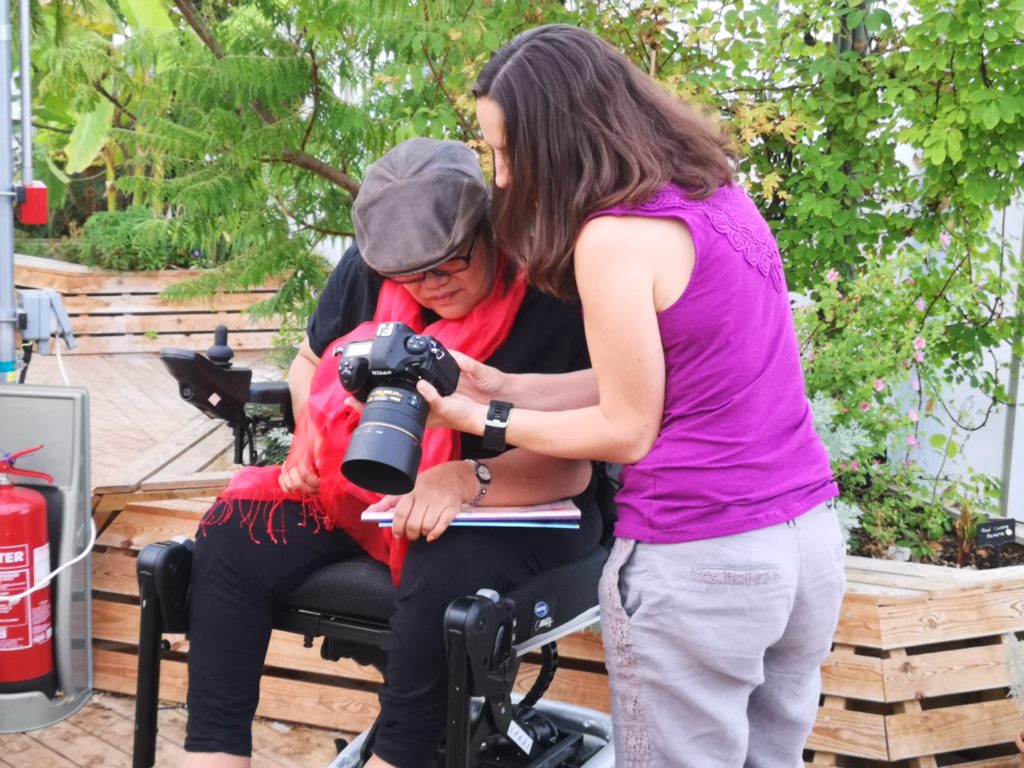 photo of standing white woman showing photos on a camera to East Asian woman with cap in a wheelchair. She is wearing a red scarf.