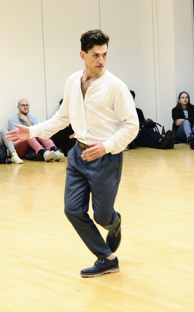 White man, in whirt shirt and dark trousers, dancing to an audience of seated audience