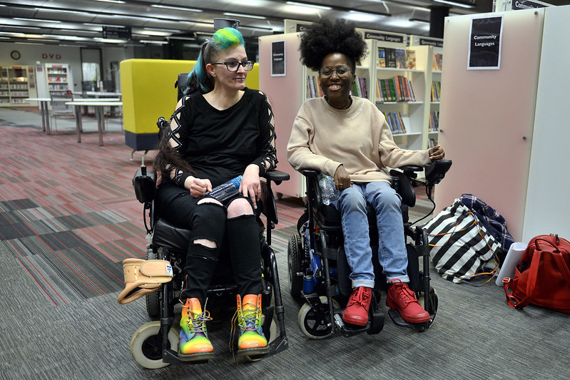 2 wheelchair users, one white with long green hair, the other a black woman. they are both smiling.
