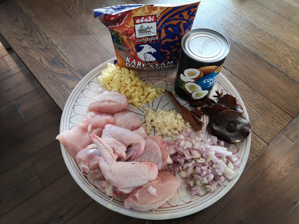 ingredients on a round plate: chicken pieces. chopped shallots, chopped garlic, chopped ginger, curry poweder in a packet (kari ayam, adabi brand), tin of coconut milk, with spices - cinnamon,  tamarind, cloves, snar anise.