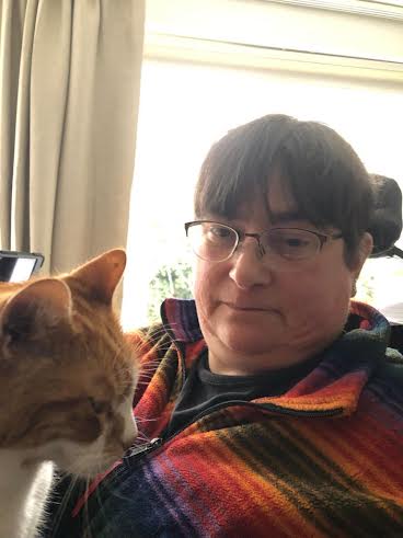 Jenny, a wheelchair user, has short hair, glasses, a stripey orange and black top.  Her cat is also in the photo.